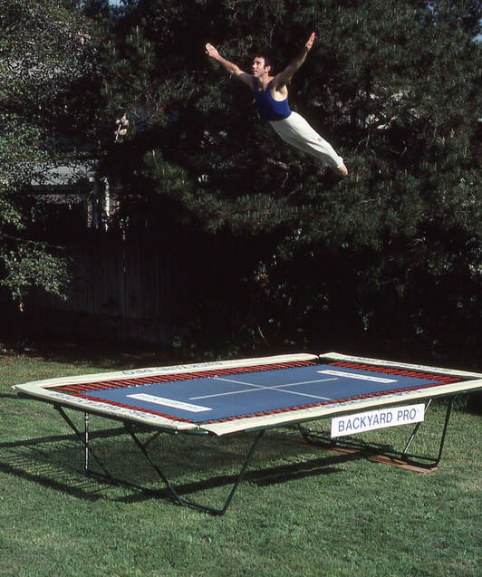 A World Trampoline Champion performs a horizontal swan dive with perfect artistic form. He is high above his own personal Backyard Pro® High-Performance Gymnastic and Sports Training Trampoline. The trampoline is outside on green grass with leafy trees in the background. The trampoline has an outdoor rated blue competition high performance string fly bed and tan safety pads. The World Trampoline Champion is wearing a navy blue gymnastic top with white gymnastic pants.