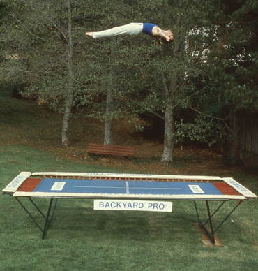 A World Trampoline Champion shows perfect form and control in a face-up horizontal position over 24 feet high on his own personal Backyard Pro® High Performance, Sports Training Trampoline. The trampoline is above ground on a green lawn with leafy trees in the background. This World Trampoline Champion is wearing white gymnastic pants and blue gymnastic top. The trampoline features our outdoor rated competition, high-performance blue string bed with white center markings. Our authoritative and expert 90 minute safety and instructional DVD is taught by this same World Trampoline Champion and is the best reviewed and most critically acclaimed  trampoline instructional & safety video ever made.