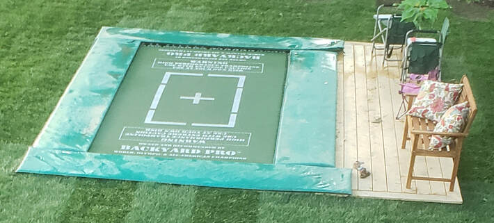 Shown is an Olympic size Backyard Pro in ground-level mode. It is surrounded by a newly turfed lawn. It features a green high performance woven string fly bed with white competition markings. It also has matching Aqua green extra wide safety frame pads. All that can be seen is the bouncing bed and the safety pads. The springs and frame our hidden for extra safety. Along one side of the trampoline is a wood observation deck with some lawn chairs. Every Backyard Pro model features a patented design that allows it to be  used either above ground or at ground level. You'll always have both options to choose from.