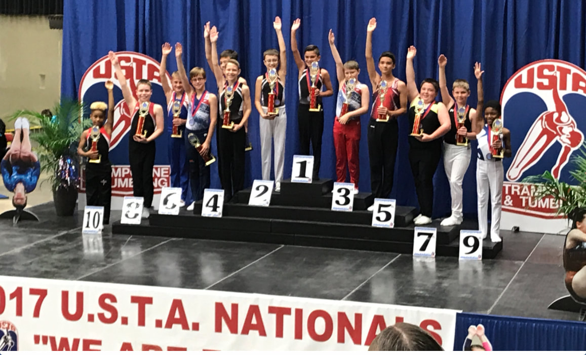 A group of about 10 boys are standing on the black finalists platform for the 10 to 12 year-old age group at the United States Trampoline Association National Championships. The boys are all wearing gymnastic tops and gymnastics pants with white trampoline slippers. Each boy is holding his trampoline trophy in his left hand while his right hand is raised above his head. The first place winner, Matthew Breese, is United States Trampoline Champion in the 10 to 12 year age group. Matthew Breese owns his own Backyard Pro® Sports Training Trampoline and affirms that has it has helped him win the United States Trampoline Championship.