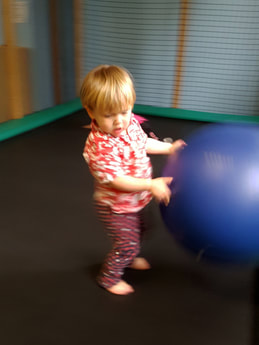 A toddler stands on the black mat of a trampoline with green safety frame pads. The toddler is playing with a large blue ball which stands shoulder height to the toddler. Trampolines make a soft forgiving play surface for toddlers.