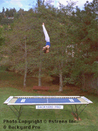 A World Trampoline Champion performs an artistic dive at great height with perfect form and control on his own personal Backyard Pro® Trampoline. The trampoline is in its above-ground mode outside on the grass with trees in the background.