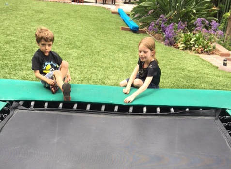Two young children about five years old are sitting on their Backyard Pro® trampoline installed at ground level. In the background are lawn, some flowers and a practice balance beam. This photo shows that the Backyard Pro® installed at ground-level is an ideal platform for even young gymnasts to play and practice on.