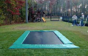 A Backyard Pro® Trampoline featuring our standard all-around outdoor black bed (also called the mat), stainless steel high performance springs, and extra-wide light green colored safety pads around the edge is installed at ground level on a green lawn. The extra-wide safety pads fully cover the springs and frame giving installation a very plush appearance. All that can be seen is the safety pads surrounding the black bed or mat. The lawn is on a beautiful upscale estate with large trees in the background. There is a wrought-iron fence around the lawn. In the background three men are standing by the fence. To the rear background is an iron gate leading to the lawn with the trampoline is placed.