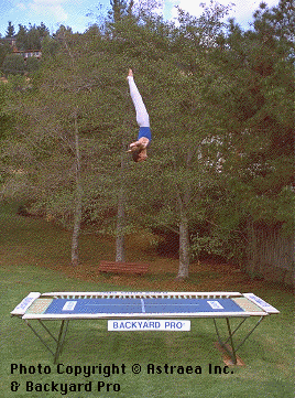 A World Trampoline Champion shows perfect form and control in a extended inverted position over 20 feet high on his own personal Backyard Pro® trampoline. The trampoline is above ground on a green lawn with leafy trees in the background. This World Trampoline Champion is wearing white gymnastic pants and blue gymnastic top. The trampoline features our outdoor rated competition, high-performance blue fly bed with white center markings. This inspirational and artistic performance is also used as the cover photo on our authoritative and expert 90 minute safety and instructional DVD which is taught by this World Trampoline Champion and is the best and most critically acclaimed trampoline instructional & safety video ever made.