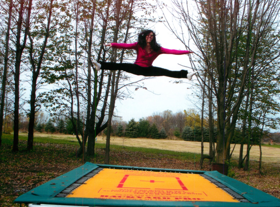 A Backyard Pro® trampoline is assembled above ground with our competition high visibility yellow string fly bed with red markings and our standard width green safety pads. Our standard pads fully cover the frame and most of the extended spring but leave about 2 inches of the spring next to the bed exposed for better performance. A girl gymnast is performing a straddle splits jump about 10 feet high. She is wearing a red top, black slacks and white socks. Her trampoline is standing on a leafy lawn with trees in the background, an open field beyond the trees and a dense stand of evergreens in the far background beyond the field.