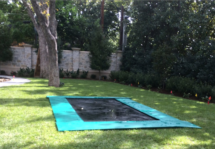A ground level Backyard Pro® Trampoline features our extra wide safety pads which we recommend for ground-level installation only. These extra wide pads fully cover the frame and springs and also extend out beyond the outside edge of the frame to overlap about another 10 to 12 inches onto the surrounding lawn to more fully cover the edge of the excavation. All that can be seen is our standard all-around outdoor black bed (also called the mat) and the extra wide pads which fully cover the high performance stainless steel springs and frame. This makes a more plush, attractive and safer installation. The surrounding lawn is beautiful and upscale with large trees and a hand built stone wall in the background with colorful plantings along the stone wall.