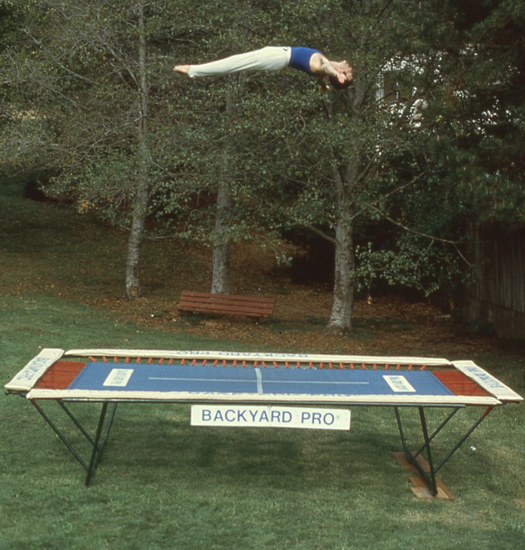 A World Trampoline Champion shows perfect form and control in a face up horizontal position over 24 feet high on his own personal Backyard Pro® trampoline. The trampoline is above ground on a green lawn with leafy trees in the background. This World Trampoline Champion is wearing white gymnastic pants and blue gymnastic top. The trampoline features our outdoor rated competition, high-performance blue string bed with white center markings. Our authoritative and expert 90 minute safety and instructional DVD is taught by this same World Trampoline Champion and is the best reviewed and most critically acclaimed  trampoline instructional & safety video ever made.