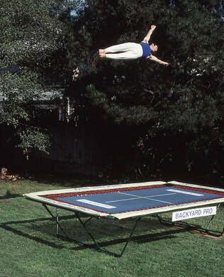 A World Trampoline Champion shows perfect form and control in a face forward sideways horizontal position over 24 feet high on his own personal Backyard Pro® trampoline. The trampoline is above ground on a green lawn with leafy trees in the background and parts of  a home peeking through the trees. This World Trampoline Champion is wearing white gymnastic pants and blue gymnastic top. The trampoline features our outdoor rated competition, high-performance blue string bed with white center markings. Our authoritative and expert 90 minute safety and instructional DVD is taught by this same World Trampoline Champion and is the best reviewed and most critically acclaimed  trampoline instructional & safety video ever made.