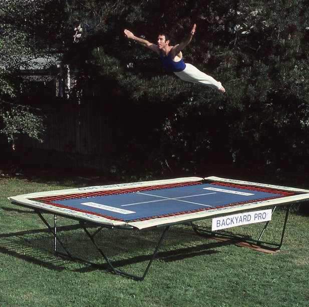 A World Trampoline Champion performs a horizontal swan dive with perfect artistic form. He is high above his own personal Backyard Pro® High-Performance Gymnastic And Sports Training Trampoline. The trampoline is outside on green grass with leafy trees in the background. The trampoline has an outdoor rated blue competition high performance string fly bed and tan safety pads. The World Trampoline Champion is wearing a navy blue gymnastic top with white gymnastic pants.