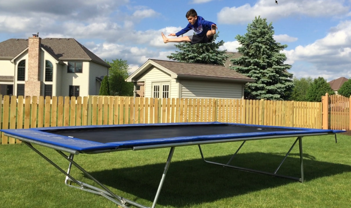 United States 10 to 12 year old age group Trampoline Champion and Backyard Pro® Sports Training Trampoline owner Matthew Breeze performs a straddle splits jump 12 feet high above his above-ground Backyard Pro trampoline. He is wearing a blue hooded sweatshirt with with dark brown shorts. His Backyard Pro® trampoline has a black all around outdoor standard bad with blue wide safety frame pads. His Backyard Pro® Trampoline stands on a green lawn. The sky is blue with a few picturesque clouds. A brown stained cedar picket fence is behind the trampoline. Behind the fence is a neighbor's light brown house with Evergreen trees in the background of the house.