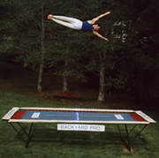 A World Trampoline Champion shows perfect form and control in a face forward sideways horizontal position over 20 feet high on his own personal Backyard Pro® trampoline. The trampoline is above ground on a green lawn with leafy trees in the background. This World Trampoline Champion is wearing white gymnastic pants and blue gymnastic top. The trampoline features our outdoor rated competition, high-performance blue string bed with white center markings. Our authoritative and expert 90 minute safety and instructional DVD is taught by this same World Trampoline Champion and is the best reviewed and most critically acclaimed  trampoline instructional & safety video ever made.
