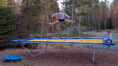 A Backyard Pro® trampoline with a yellow competition bed and high performance stainless steel springs sits above ground in a clearing in a wooded area. An eight-year-old girl is doing a splits jump with an even younger boy observing from the from the built-in blue padded observation deck of the trampoline.