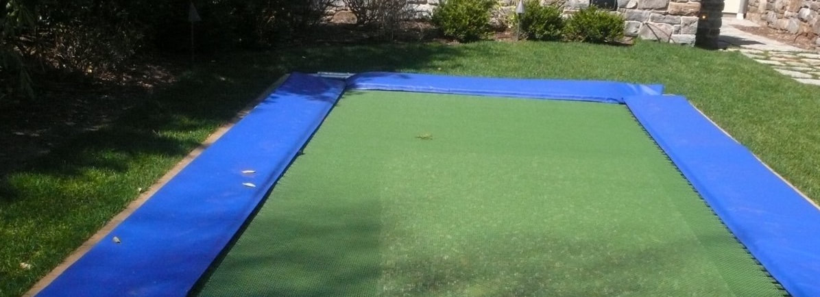 A ground-level Backyard Pro® features optional wide blue safety pads. Our optional wide safety pads fully cover the frame and springs with no part of the springs left exposed. It also has a green, high-performance outdoor rated competition string fly bad. The trampoline it is surrounded by a plush green lawn. A stone wall is in the far background with some plantings along the wall.